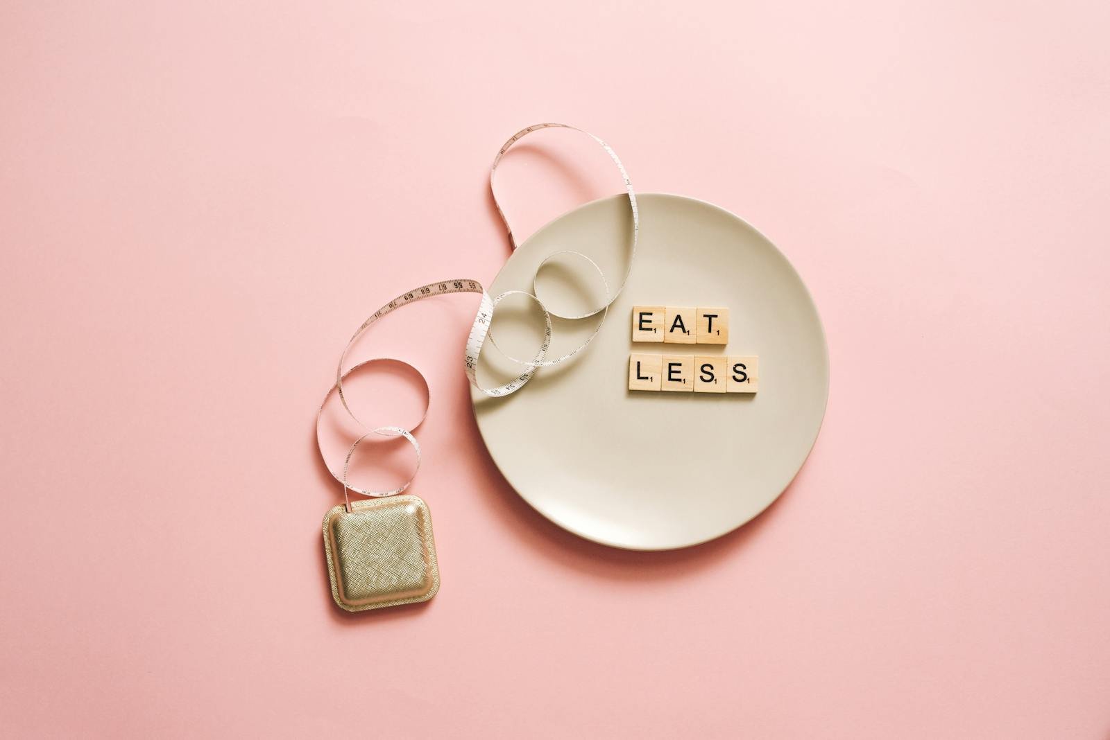 A motivational image featuring a plate with scrabble tiles spelling 'Eat Less' on a pink background.