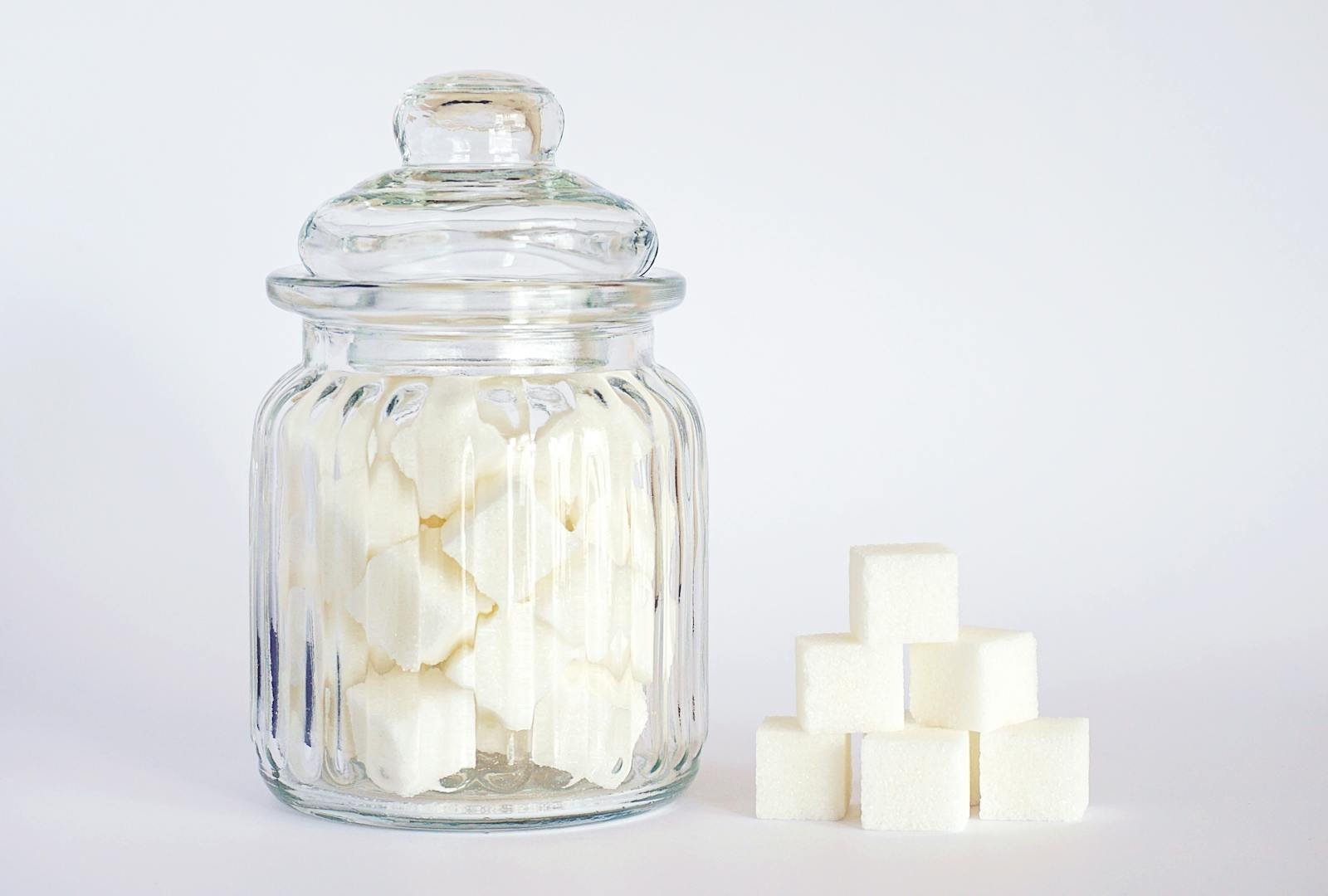 A clear glass jar filled with sugar cubes next to stacked cubes on a white background.