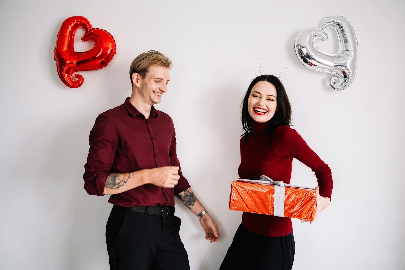 A joyful couple exchanging gifts surrounded by heart-shaped balloons, embracing a romantic celebration.