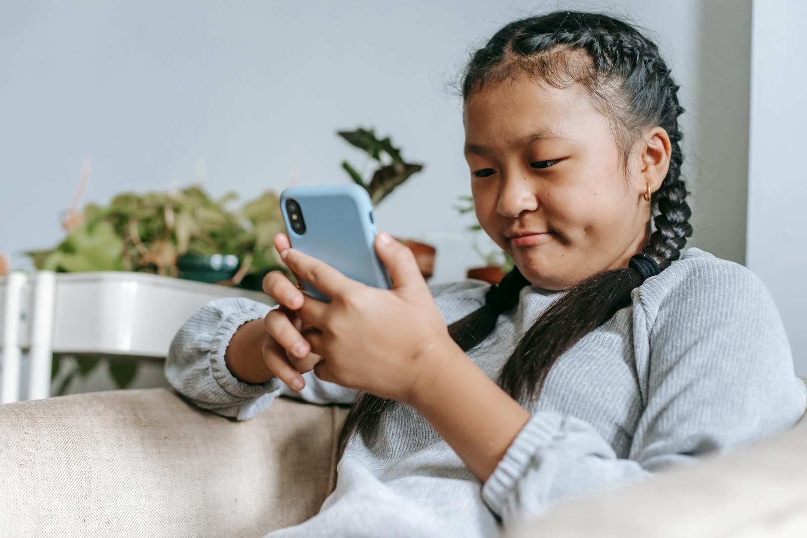 A young girl with braided hair using a smartphone on a couch, illustrating digital pastime.