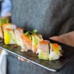 Colorful sushi rolls with salmon, avocado, and roe served on a ceramic plate by a chef in a restaurant.