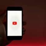 A hand holding a smartphone displaying the YouTube app against a red background.